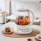 A BUYDEEM electric beverage maker with a white base sits on a woven placemat on a white table, filled with orange and dark herbal tea. A matching glass cup with tea is on the left, and wooden spoons and a plate are on the right, set against a softly blurred modern interior background.