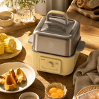A countertop electric food steamer with a stainless steel upper chamber and cream-colored base, styled on a wooden table with breakfast items including corn, pumpkin, eggs, toast, and a mug in warm natural light.